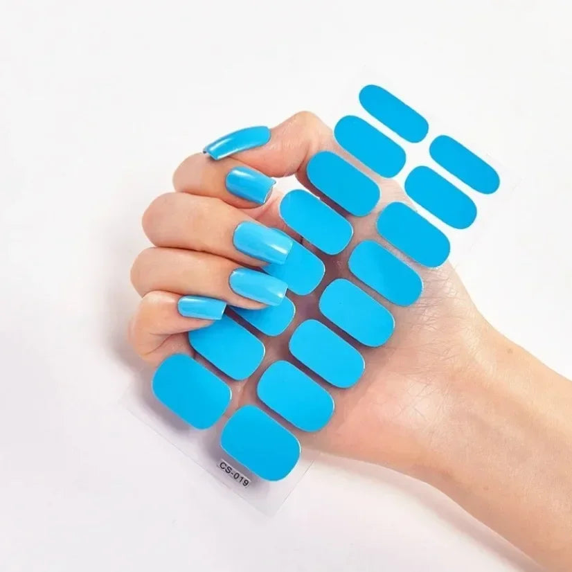Long, light blue press-on nails held in a hand next to a sheet of matching blue nail wraps.