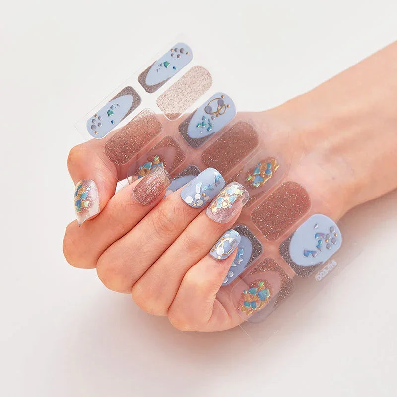 Currant™ nail wraps on a hand, showcasing blue, glittery designs with floral and geometric patterns.