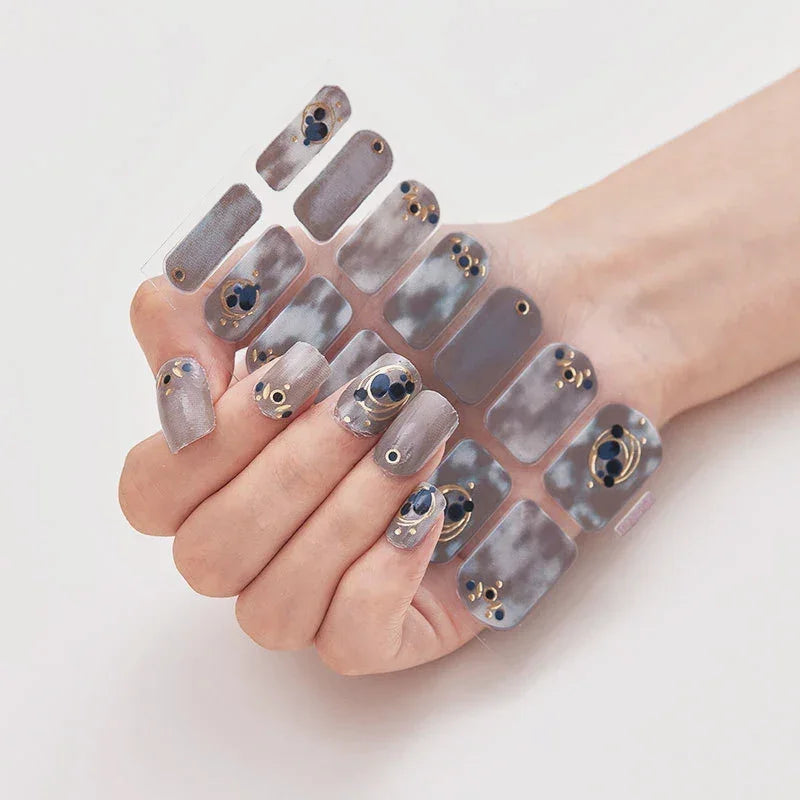 Currant™ press-on nails displayed on a hand, featuring gray and blue marble patterns with gold accents and gems.
